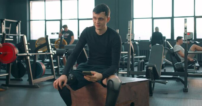 Disabled young man with prosthetic amputee leg using smart phone while resting at gym, send message, sms. e-mail, chat with friends browse, surf the Net Slow motion gadget guy look at screen of phone