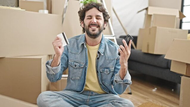 Young hispanic man shopping with smartphone and credit card sitting on floor at new home