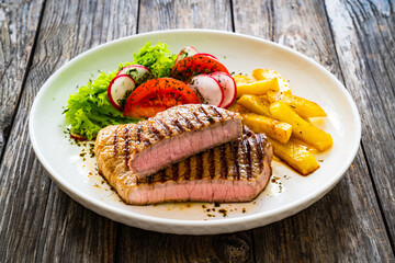 Grilled beef steaks with fresh vegetable salad and French fries on wooden table
