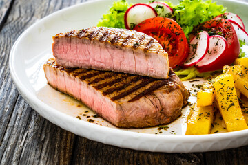 Grilled beef steaks with fresh vegetable salad and French fries on wooden table
