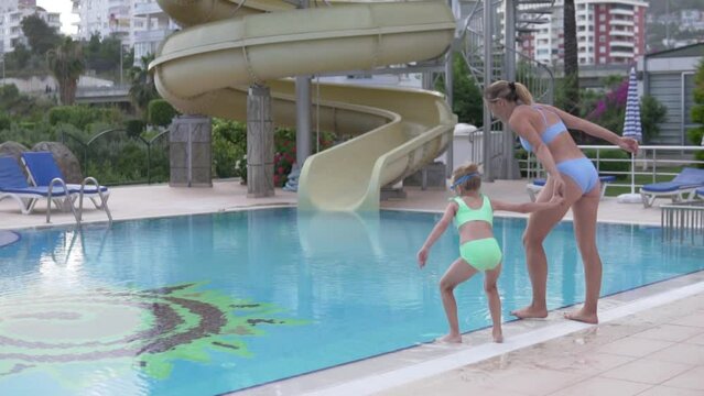 Mom and little daughter jump into the pool in the summer, holding hands.