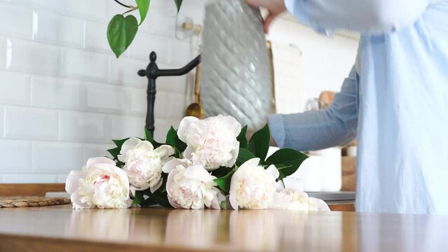 Woman bringing beautiful white peony flowers to the kitchen and adding water to vase