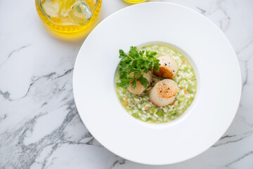 White plate with pea risotto and seared sea scallops, horizontal shot on a white marble background, high angle view