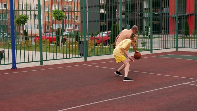 A happy father and a teenage daughter play basketball outdoors on the court, in the courtyard of a multi-storey building.