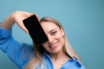 smiling stylish blond girl holding smartphone screen with mockup for web page near her face on blue...