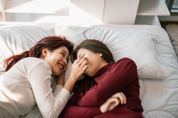 Fototapeta na wymiar Asian lesbian partners smile, relax, laugh, affectionately pinch and kiss each other's cheeks while lying on the bed. A combination of personal charm, a good sense of humor, and a romantic moment.