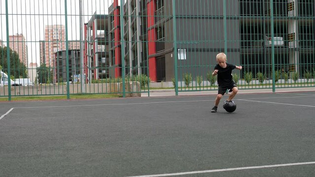A little boy trains with a soccer ball on a sports court against the background of a multi-storey building.