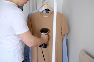 A young man holds the garment steamer in his hand and smoothes the t-shirt after washing and drying - obrazy, fototapety, plakaty
