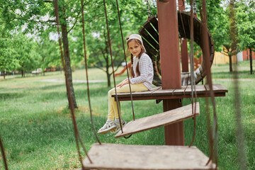 Little tired girl wearing casual attire and cap sitting on rope swings at sunny playground having fun resting having break of active playing.