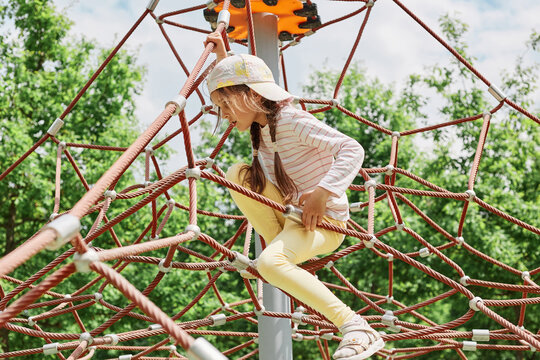 Little girl with pigtails and cap playing in rope polyhedron climb at playground outdoor having fun in camp or in park kid's sport activity in beautiful nature.