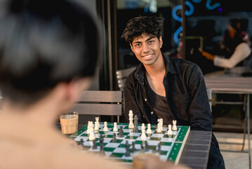 An asian guy in a black jacket and black shirt is smiling at the camera, while playing chess with...