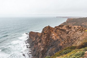 Adventurous man standing on the edge of a cliff enjoys the view of the Atlantic coast in the Odemira region of southwestern Portugal. Wandering the Rota Vicentina