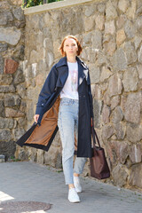 Elegant middle aged woman walks city street wearing trench coat, jeans and sneakers