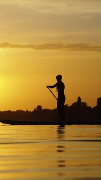 Vertical Screen: Silhouette of woman paddling on SUP board in slow motion, sunset over river. Pan shot sporty female doing sports in beautiful landscape. Healthy lifestyle. Concept of fitness