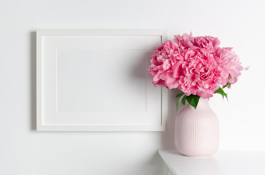 Blank horizontal frame mockup on white wall with pink peony flowers for artwork presentation, empty frame with copy space