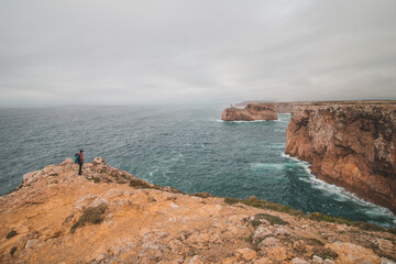Black-haired adventurer stands at the end of Cape Cabo de Sao Vicente in the southwest of Portugal in the Algarve region. Man is enjoying his freedom. Wandering of Fisherman Trail