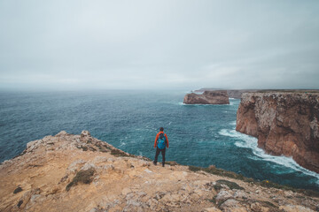 Black-haired adventurer stands at the end of Cape Cabo de Sao Vicente in the southwest of Portugal in the Algarve region. Man is enjoying his freedom. Wandering of Fisherman Trail