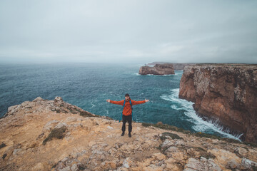 Fototapeta premium Black-haired adventurer stands at the end of Cape Cabo de Sao Vicente in the southwest of Portugal in the Algarve region. Man is enjoying his freedom. Wandering of Fisherman Trail