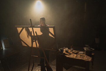 Female artist works on abstract oil painting, creates modern masterpiece. Dark creative studio with large canvas illuminated, painter workplace.