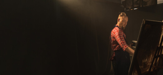 Talented male artist creates modern masterpiece of the painting using paint brush. Working in artist workshop. Dark creative studio with large canvas. Side view