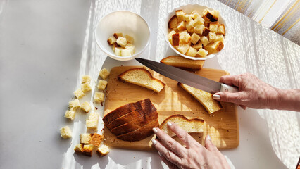 Fototapeta premium Homemade cooking and cutting of slices of diced dry white rye bread on cutting board and white plate. Hand of woman with Crackers for food, healthy eating on table in sunny day, top view