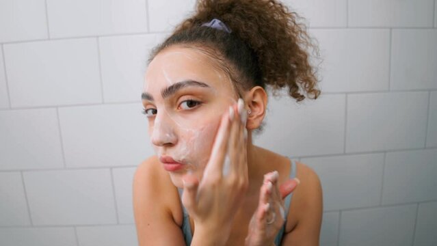 girl performing daily facial skincare procedure. Young woman is washing applying foam cleanser on face. looking at camera, slow motion