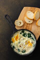 Healthy breakfasts or dinners, fried eggs with cheese, spinach leaves and coconut milk in a cast-iron pan, sliced baguette on a cutting board on a brown background