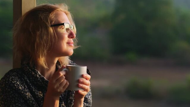 Mid adult woman wearing glasses drinking hot coffee at sunrise outdoor. Pleased female blonde enjoys beautiful morning. Happy caucasian woman portrait on the blurred nature background.