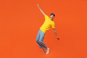 Fototapeta na wymiar Full body young man wear pyjamas jam sleep eye mask rest relax at home stand on toes with outstretched hands lean back isolated on plain orange background studio portrait. Good mood night nap concept.