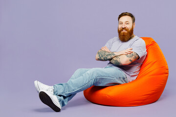 Full body young redhead bearded man wear violet t-shirt casual clothes sit in bag chair hold hands crossed folded isolated on plain pastel light purple background studio portrait. Lifestyle concept.