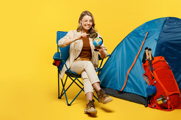 Obraz premium Full body young woman sit near bag with stuff tent hold Earth world globe magnifier isolated on plain yellow background Tourist active lifestyle walk spare time Hiking trek rest travel trip concept
