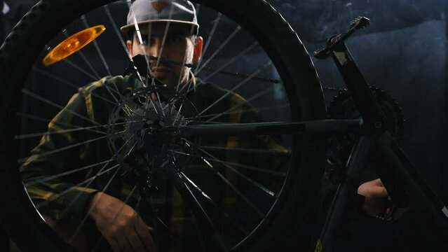 A cyclist repairs a bicycle in a dark smoke-filled garage. A man in a shirt and baseball cap spins the wheel of a mountain bike and looks for faults