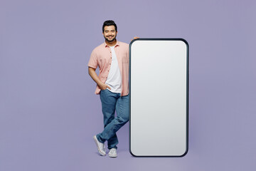 Full body smiling young Indian man he wear pink shirt white t-shirt casual clothes stand near big huge blank screen mobile cell phone smartphone with area isolated on plain pastel purple background.