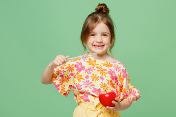 Little happy cute child kid girl 6-7 years old wearing casual clothes have fun brush teeth hold red apple isolated on plain pastel green background studio. Mother's Day love family lifestyle concept.