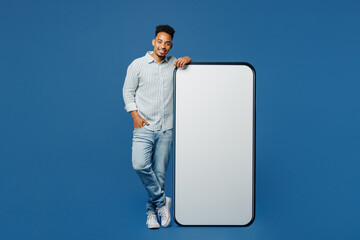 Full body fun young man of African American ethnicity wear casual clothes shirt big huge blank screen mobile cell phone smartphone with area look camera isolated on plain dark royal blue background.