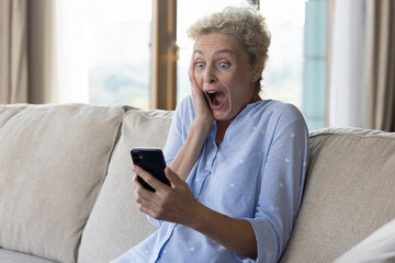 Shocked excited pretty mature woman holding mobile phone, gasping, touching face with open mouth and wide eyes in surprise, shouting for joy, celebrating win, success, victory