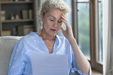 Shocked senior elder woman holding paper legal document, sitting on home sofa, touching head with...