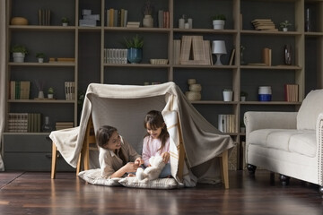 Happy mom and adorable little child girl having fun in play tent, resting on heating floor in handmade toy fort from chairs and blanket, talking, chatting, laughing - obrazy, fototapety, plakaty