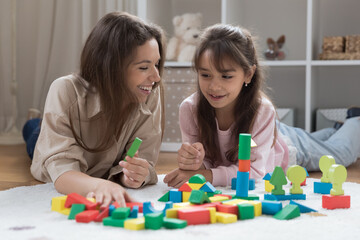 Happy cheerful young mother and adorable preschool kid girl resting at colorful cubes in playroom, chatting, laughing, having fun, playing toy building blocks on warm floor