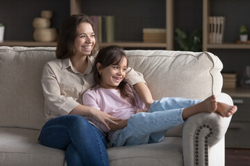 Positive young mom and dreamy sweet daughter kid looking away in happy thoughts, hugging on sofa, resting on home couch together, smiling, laughing, dreaming, thinking on future plans