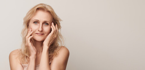 Beautiful blond middle aged woman smiling looking camera touching face portrait. Elegant mature...
