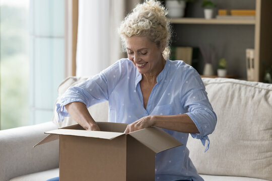 Cheerful excited customer woman getting parcel, opening cardboard box at home, checking package, getting purchase from internet store, courier delivery service, unpacking paper container