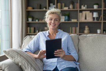 Happy pretty mature retired woman holding digital tablet computer, looking at camera with toothy smile, sitting on sofa at home. Portrait of senior customer satisfied with domestic Internet service