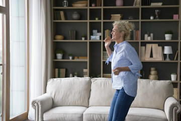 Happy beautiful senior woman dancing at home, laughing, smiling, having fun in modern spacious cozy living room, enjoying movement, motion, activity, leisure, good health, healthy joints