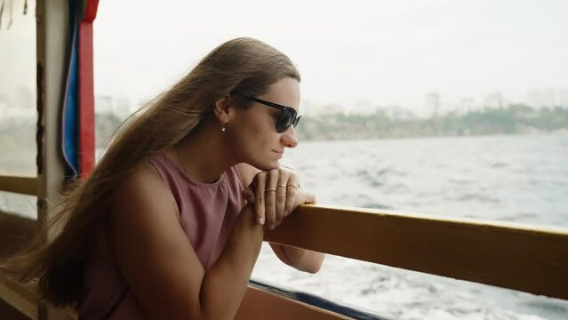 A young woman is lost in thought as she travels on the ship, gazing out at the sea with a city in the background. slow motion.