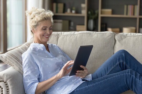 Cheerful pretty mature woman using online application on tablet computer at home, resting on couch, enjoying domestic Internet connection, communication, technology, leisure with digital gadget - Powered by Adobe