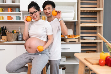 Asia pregnant mother with husband holding orange fruit in kitchen at home	