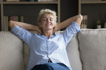 Positive calm mature senior woman resting on couch with closed eyes, relaxing, stretching body, leaning on back with open arms, enjoying silent break, leisure at home, meditating breathing fresh air