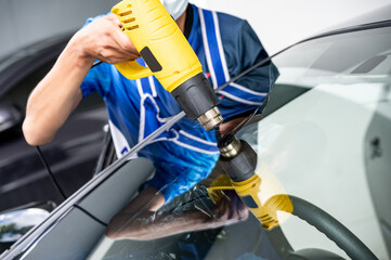 Auto specialist worker hand blowing hot air dryer or hairdryer removing old car window film tint and installing the new one. Car front windscreen film removal and tinting installation