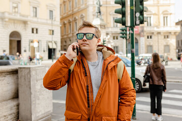Fototapeta premium Happy young man with smartphone calling on city street. Technology, communication and people concept. Handsome man speak phone wear jacket on urban background